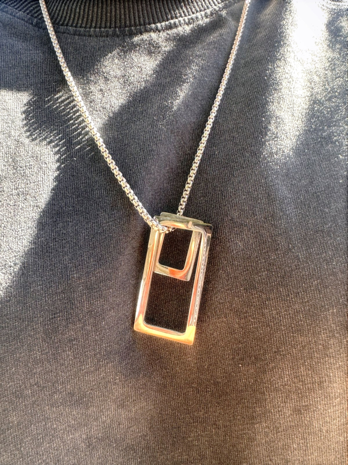 Cadena veneciana con dije rectangular y cuadrado puesta en camisa negra y brillando por la luz del solsv